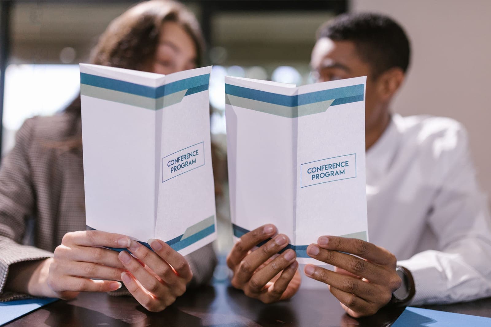 Two people in professional clothing holding and reviewing white conference program booklets with blue design elements.