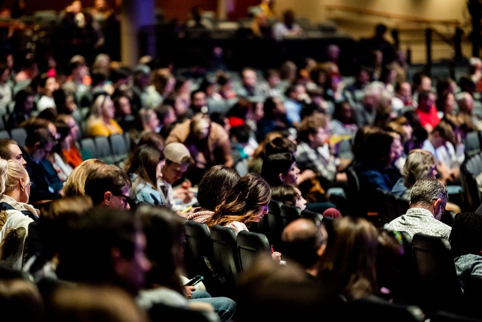 Dense crowd of conference or event attendees seated in an auditorium with stage lighting visible in background.
