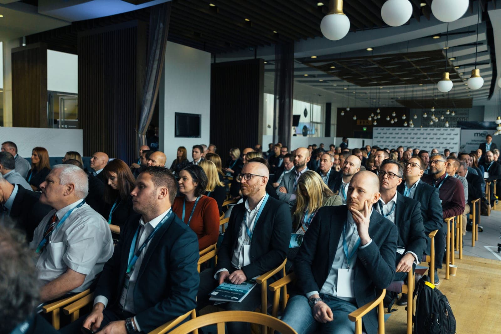 Rows of seated conference attendees wearing lanyards and badges in a modern venue with pendant lighting.