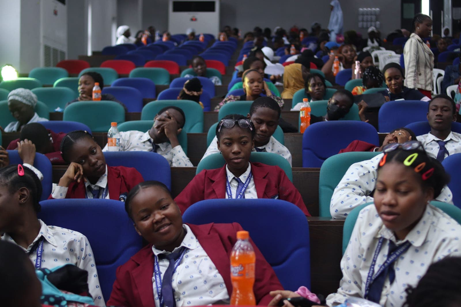 Diverse audience members seated in blue and teal chairs at a conference, wearing identification badges and casual attire.