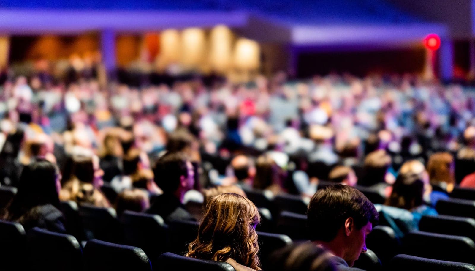 View of large conference crowd from stage perspective, with audience members seated facing forward toward a presentation area with purple stage lighting.