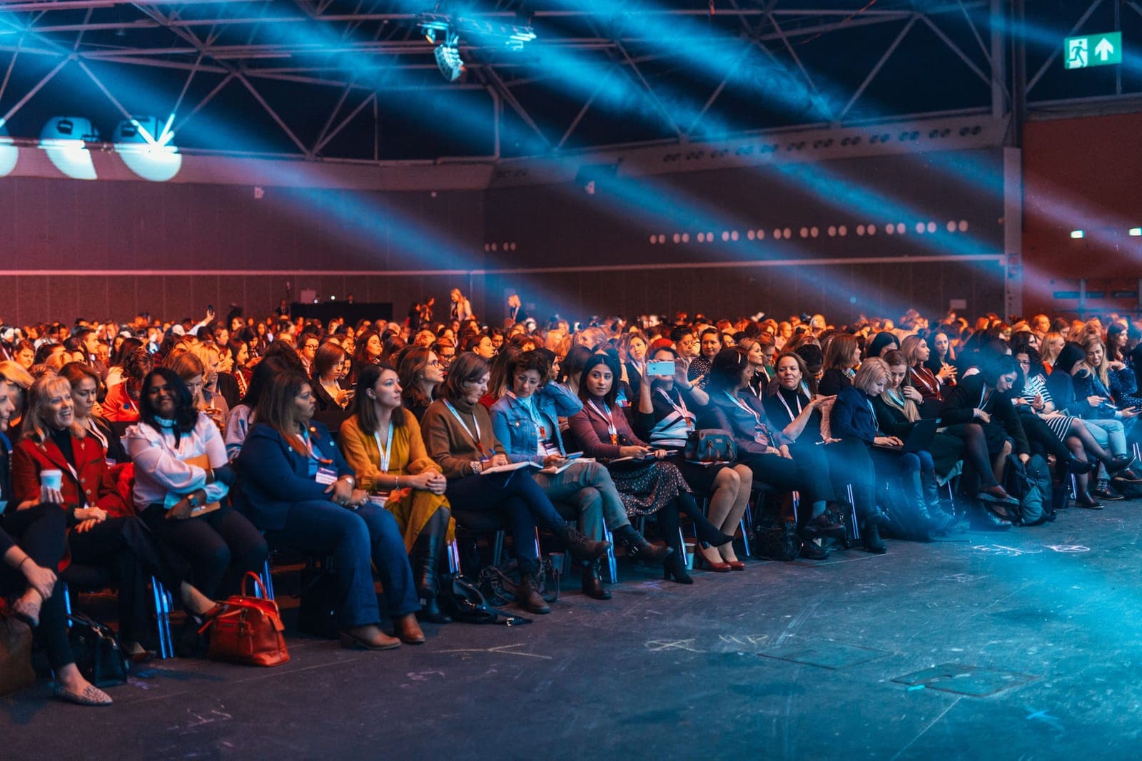 Large audience seated in rows facing a stage with blue lighting and a presenter on stage.