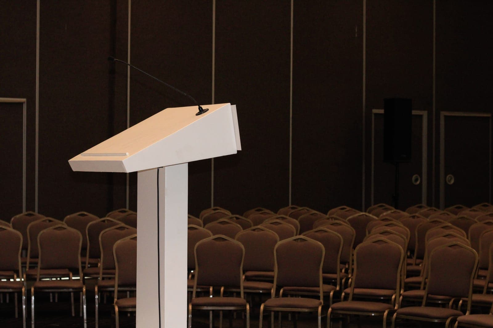 Empty white podium stands in an auditorium with rows of brown chairs, ready for a speaker.
