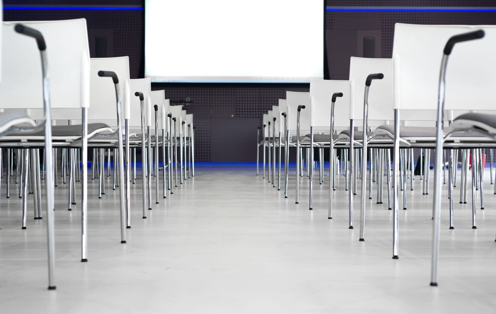 Rows of empty white and metal chairs arranged in a conference venue with a dark stage and blue lighting ahead.