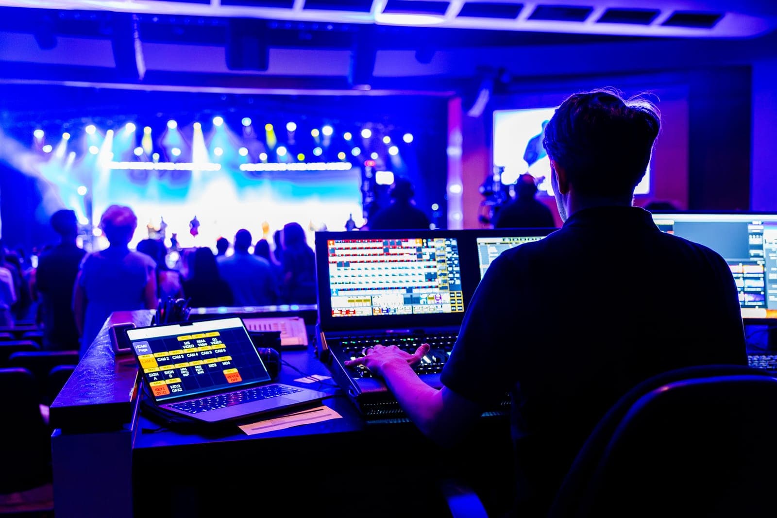 Audio-visual technician operating mixing console and multiple screens at an event, silhouetted against stage lighting and large crowd.