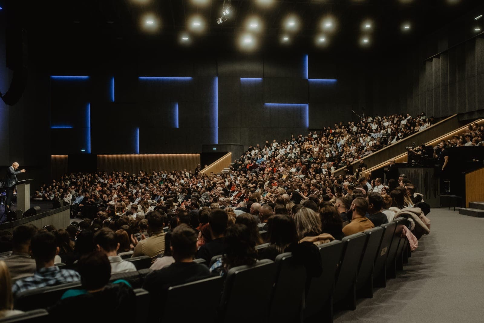 Large auditorium filled with hundreds of attendees seated in rows facing a stage with blue and yellow lighting, speaker visible on stage.