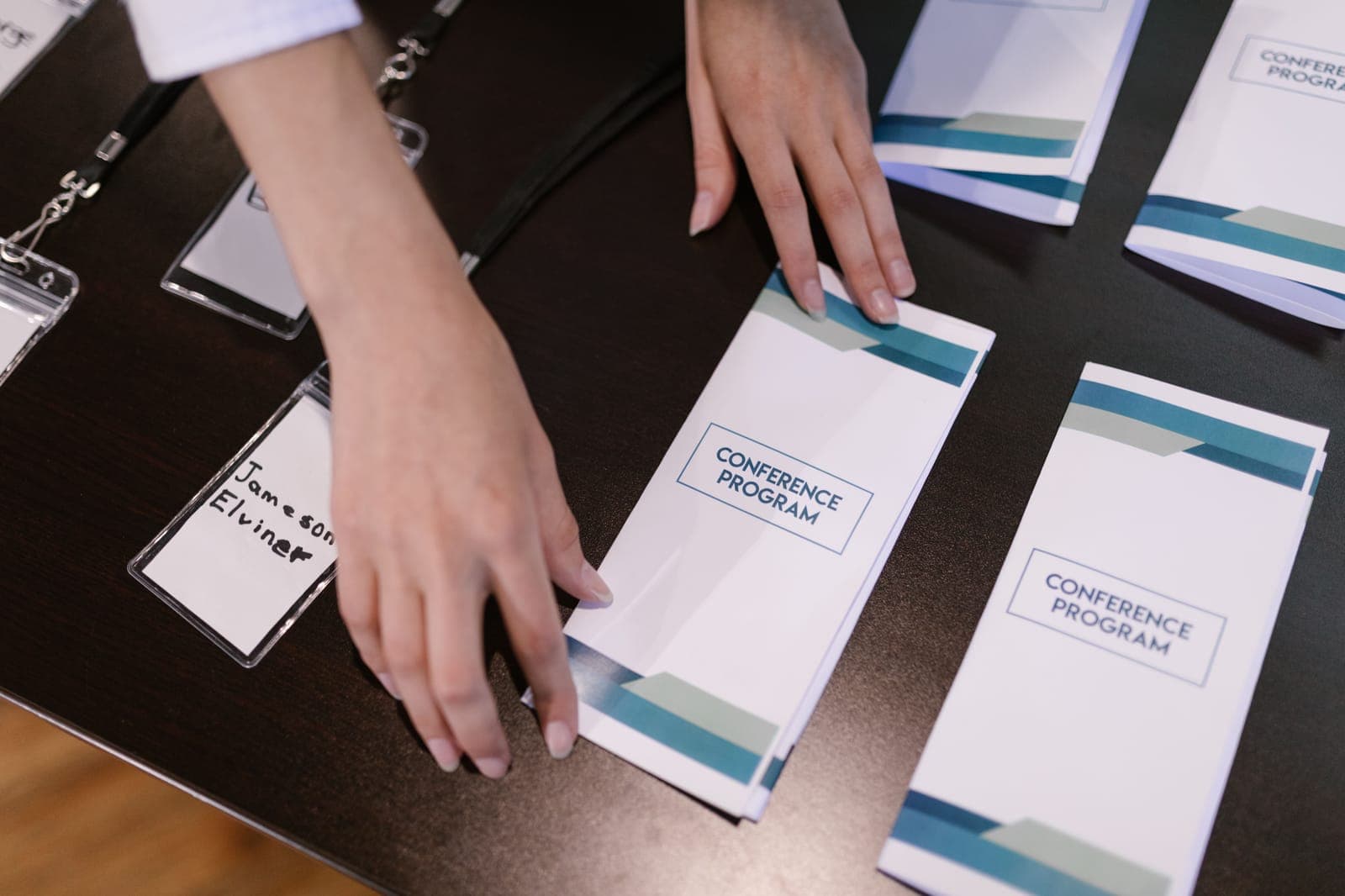 Person's hand arranging multiple white conference program booklets on a dark table surface.