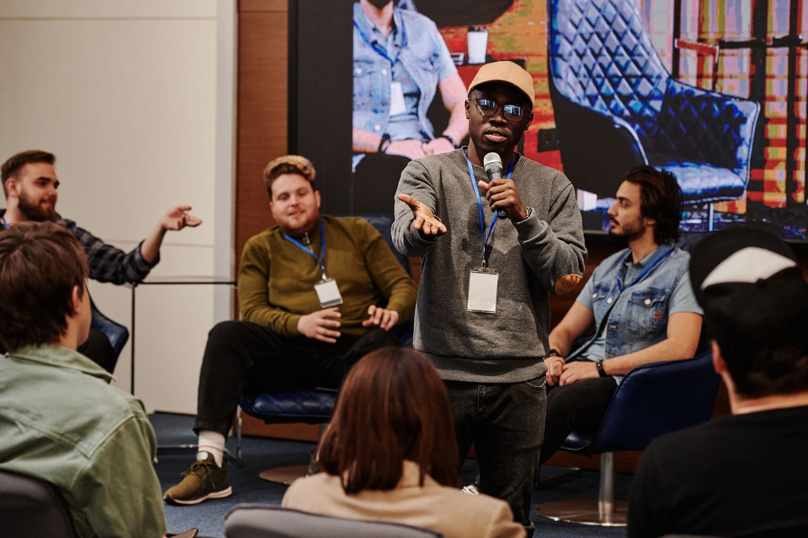 Panel moderator with microphone speaking to an audience, with other panelists seated nearby and a screen visible in the background.