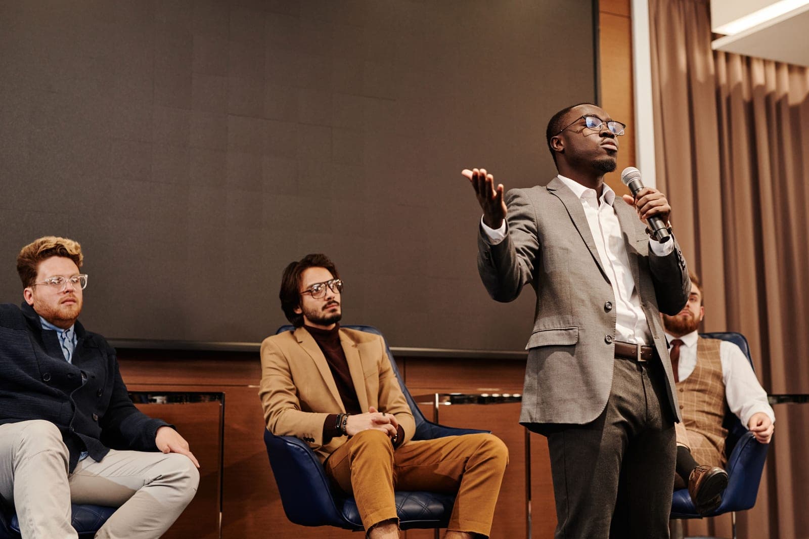 A speaker standing with a microphone addressing the room while three panelists listen from seats on stage.
