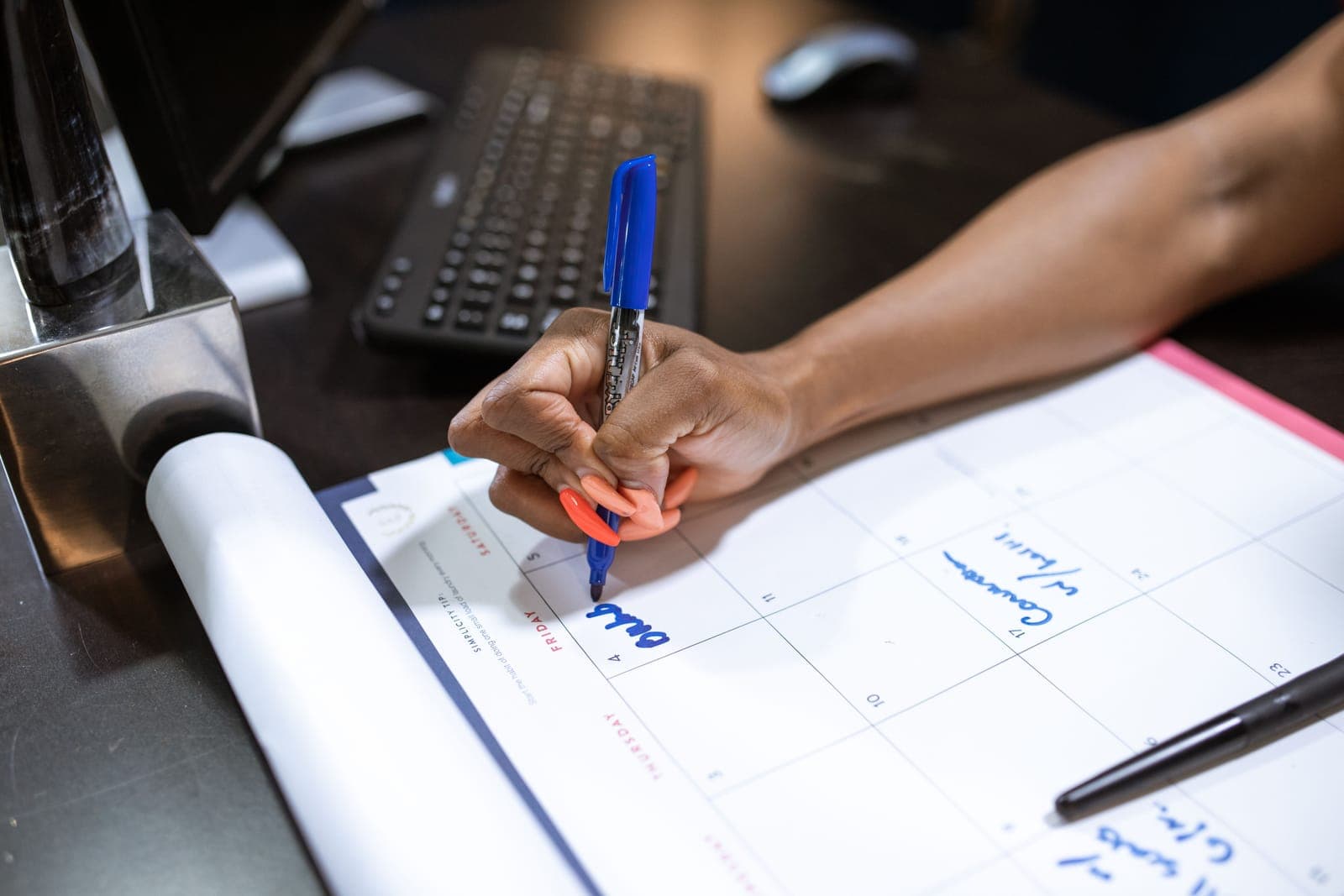 Hands of a person holding a pen while writing on a calendar or schedule at a desk with laptop in background.