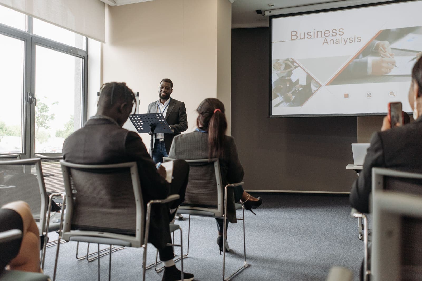 Man in business attire presenting to seated audience members in a conference room with a projection screen behind him.