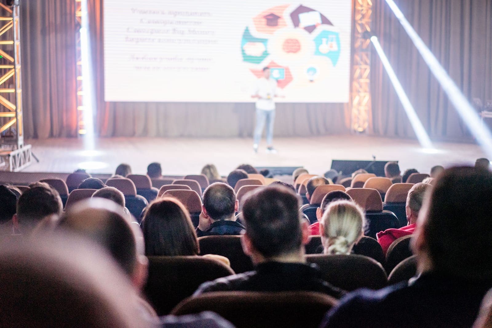 Audience members viewing a presenter on stage with a colorful projected presentation visible on screen behind the speaker.