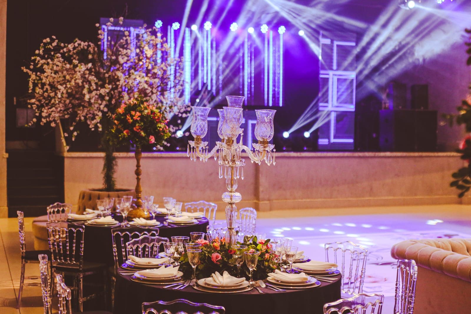 Elegantly decorated gala table in foreground with stage backdrop featuring purple lighting, geometric set design, and crystal chandelier centerpiece.
