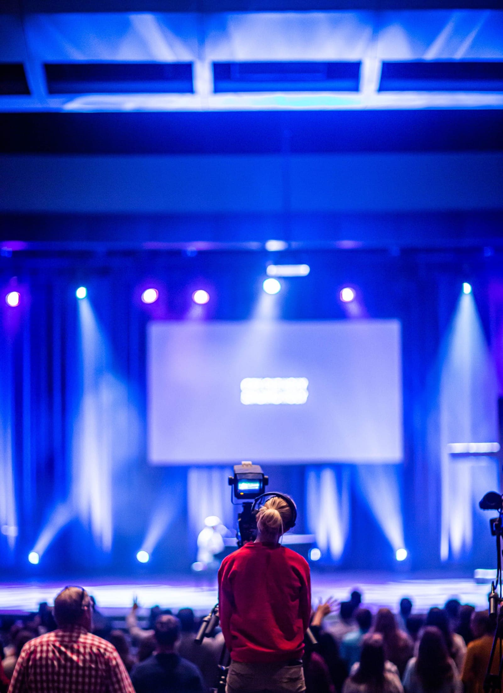 Speaker on stage with podium addressing audience, professional stage lighting with blue uplighting and projection screen visible.