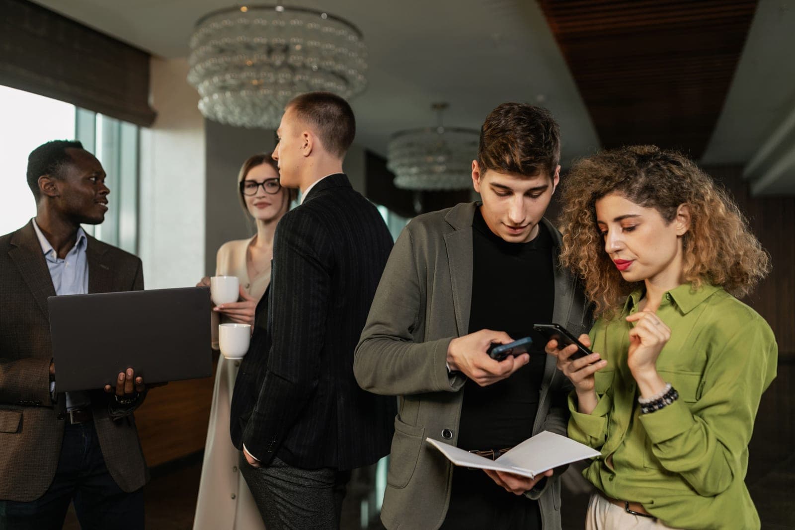 Colleagues in business clothing conferring over an open notebook, phones, and a laptop in a hotel event lobby.