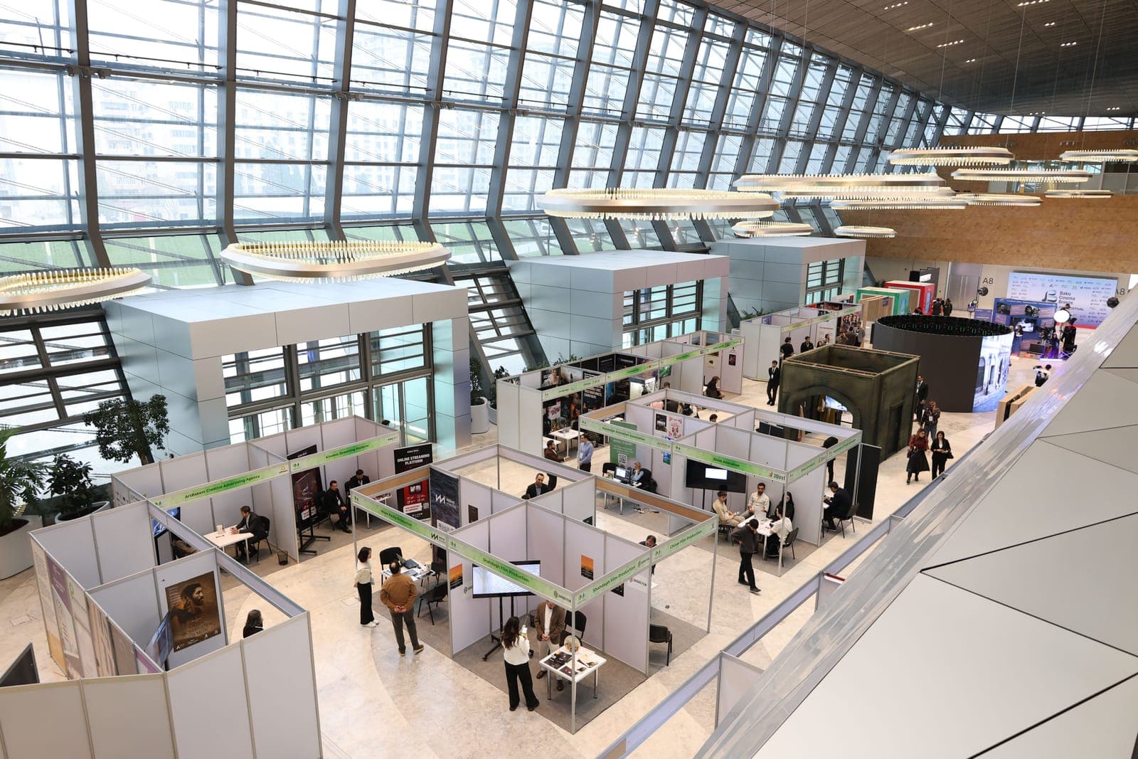 Aerial view of a large trade show or expo floor with multiple white vendor booths, attendees walking, and extensive glass ceiling.