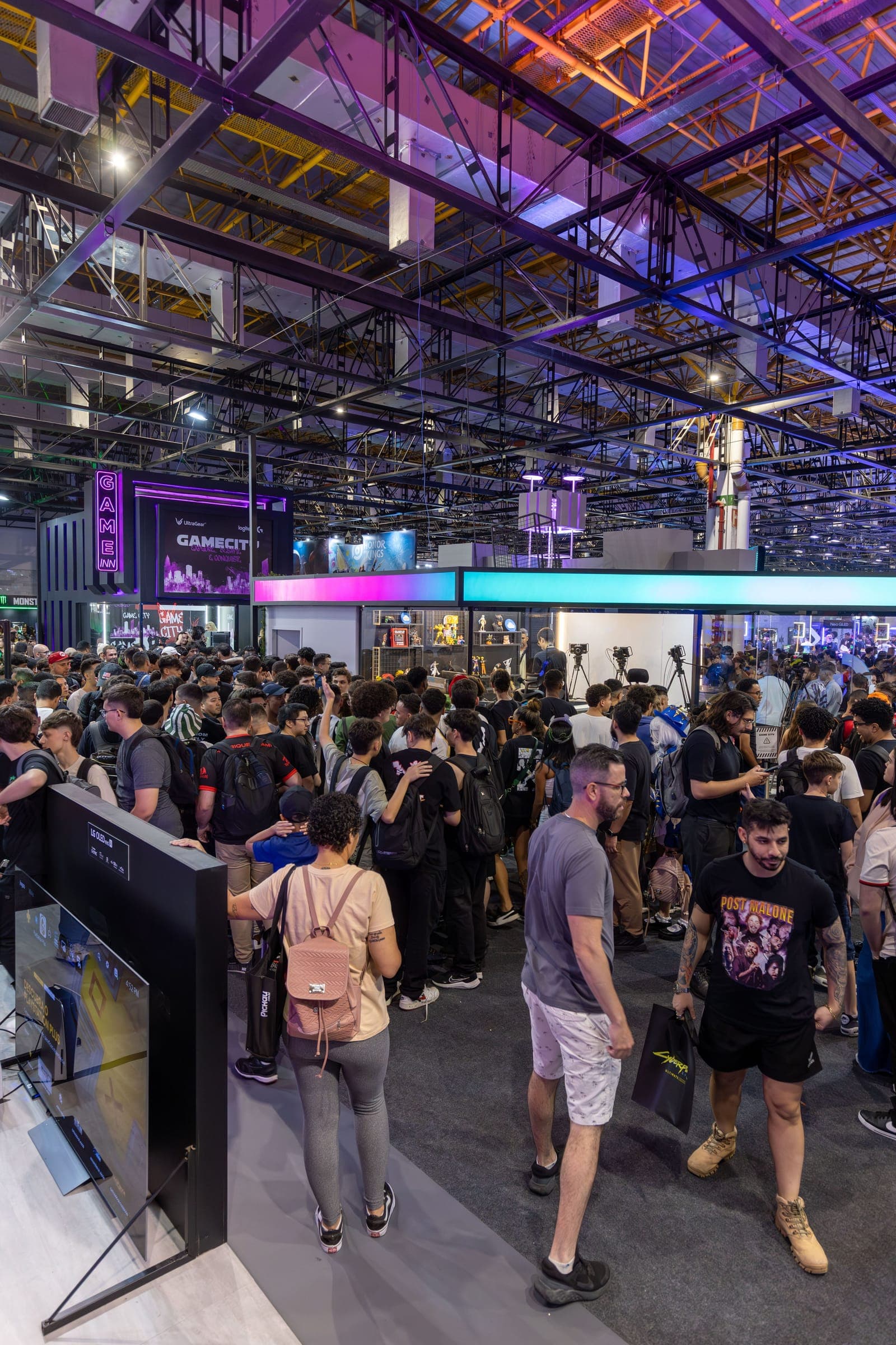 Large crowded trade show floor with multiple exhibition booths, purple and blue lighting, attendees browsing displays and vendor stations.