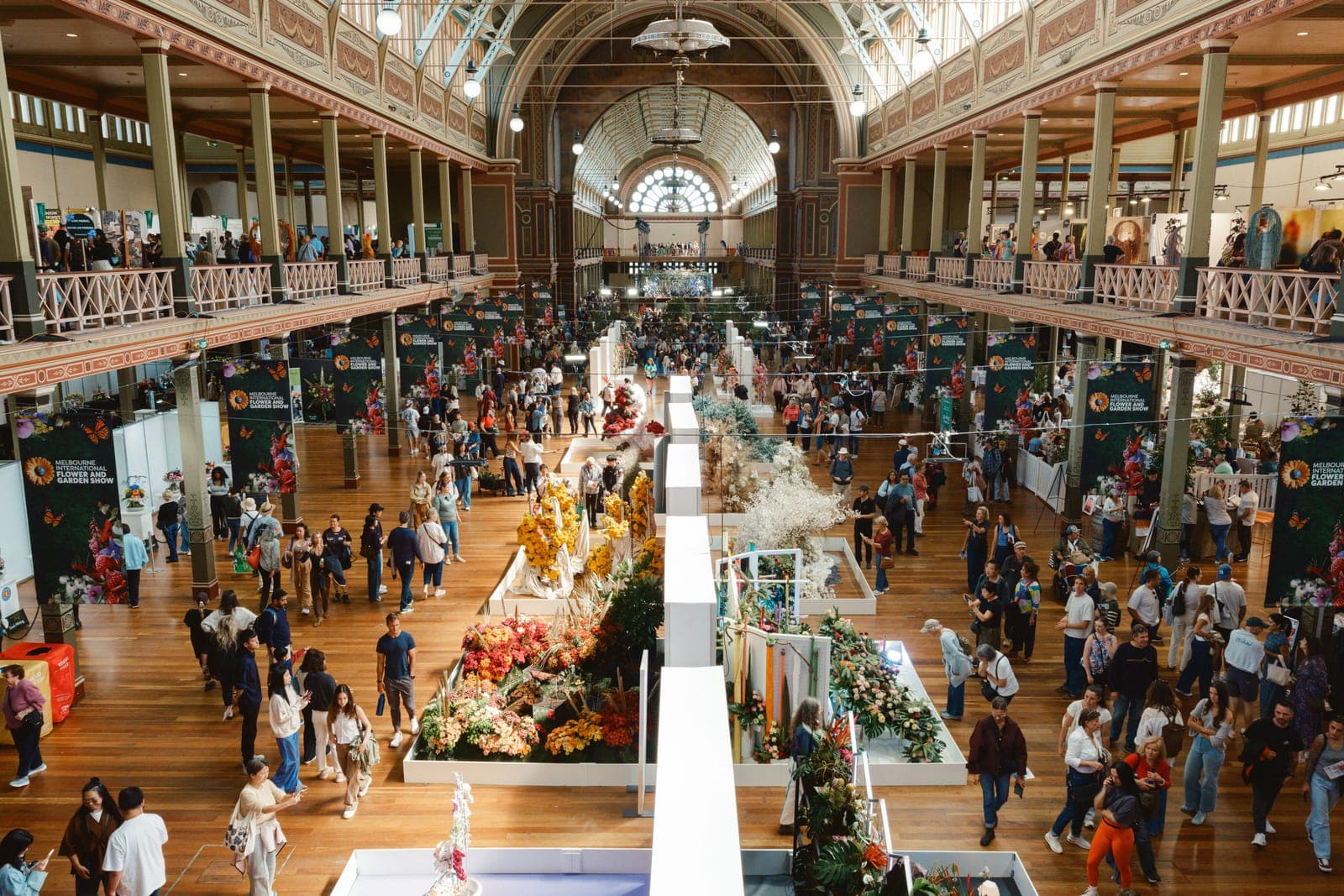 Overhead view of a bustling trade show floor in an ornate hall with vendor booths, crowds, and two-level balconies.
