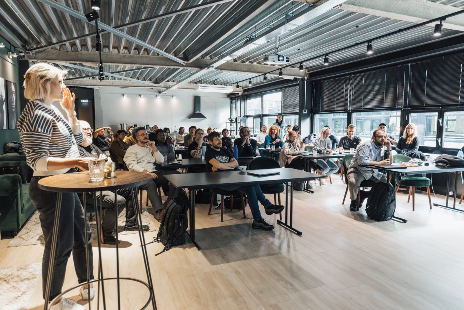 Speaker standing at podium presenting to seated attendees in a modern open office space with industrial ceiling and large windows.